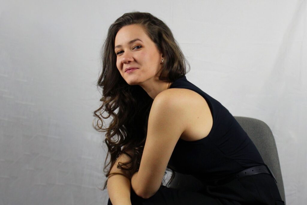 Kate, the leader of The Igniting Studio, smiling confidently with wavy brown hair and wearing a black sleeveless outfit.